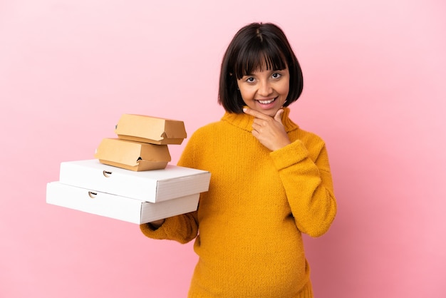 Zwangere vrouw met pizza's en hamburgers geïsoleerd op roze achtergrond gelukkig en glimlachend