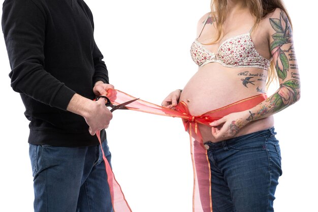 Zwangere vrouw met rood lint rond buik poseren met echtgenoot in studio op een witte achtergrond. Man snijden lint met een schaar