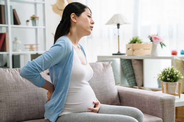 Zwangere vrouw met rugpijn in het laatste trimester van de zwangerschap. Aziatische zieke jonge vrouwelijke moeder die de taille aanraakt en een grote buikbaby binnen fronst. ongelukkige vermoeide dame die op de bank thuis rust