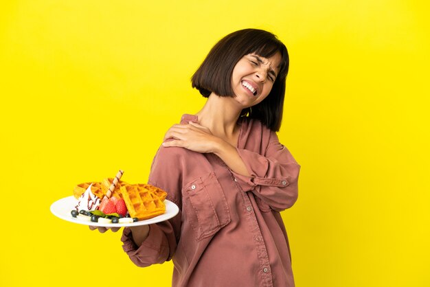 Zwangere vrouw met wafels geïsoleerd op een gele achtergrond die lijdt aan pijn in de schouder omdat ze moeite heeft gedaan