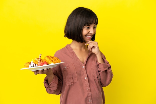 Zwangere vrouw met wafels geïsoleerd op een gele achtergrond die naar de zijkant kijkt en glimlacht