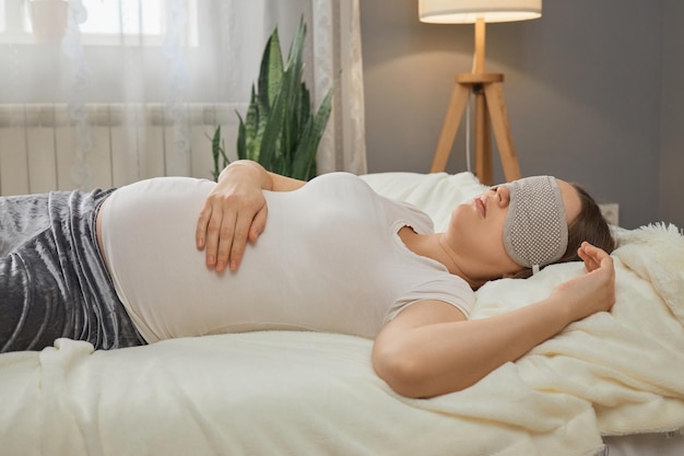 Zwangere vrouw met wit casual T-shirt en blinddoek slaapt thuis in bed in het interieur van de slaapkamer raakt haar buik aan en rust met plezier in de middag