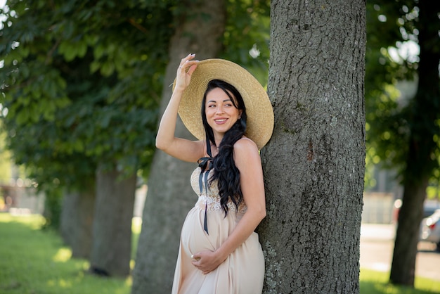 Zwangere vrouw poseren in een beige jurk met groene bomen.