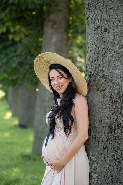 Zwangere vrouw poseren in een beige jurk op een achtergrond van groene bomen.