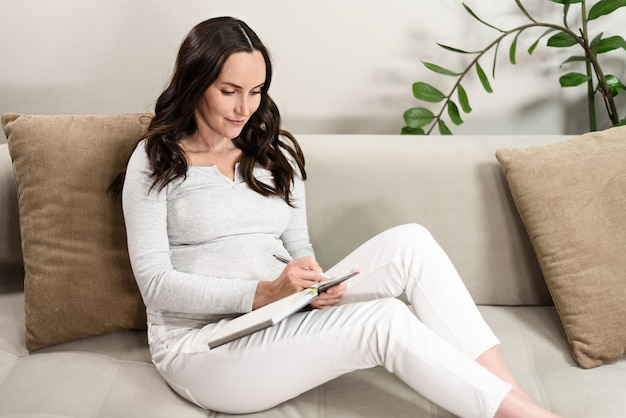 Zwangere vrouw schrijft in notitieboekje in grijze blouse zittend op de bank, maakt lijstconcept