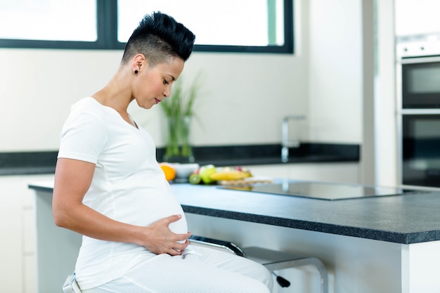 Zwangere vrouw wat betreft haar maag terwijl het zitten dichtbij keuken worktop