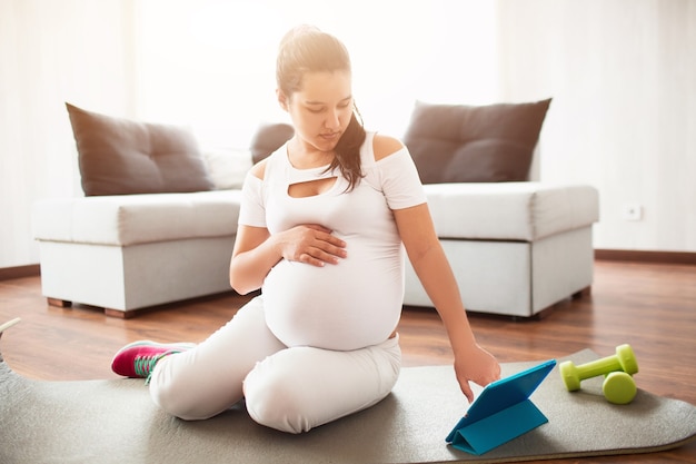 Zwangere vrouw zit op een yogamat en gebruikt een mobiele applicatie op een tablet om te sporten tijdens de zwangerschap.