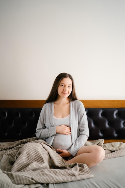 Zwangere vrouw zit op het bed en knuffelt haar buik met haar handen