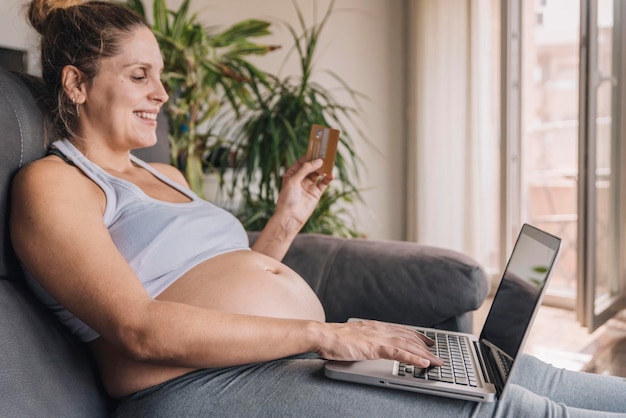 Zwangere vrouw zittend op de bank met behulp van een laptop om thuis te winkelen en te betalen met haar creditcard