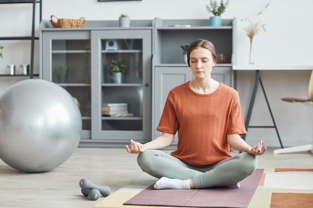 Zwangere vrouw zittend op een oefenmat in lotuspositie en mediteren met gesloten ogen in de kamer thuis