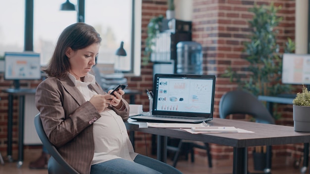 Zwangere werknemer typen op het scherm van de smartphone, bezig met zakelijk project. vrouw verwacht een kind, gebruikt een mobiele telefoon om berichten te sms'en terwijl ze in een opstartkantoor zit. persoon die aan de telefoon werkt