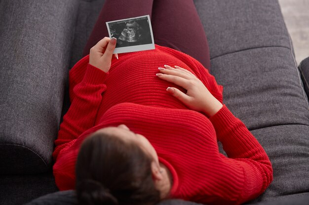 Zwangerschap, gezondheidszorg en gynaecologie concept. De mening van hierboven van zwangere vrouw houdt echobeeld, zit bij bank