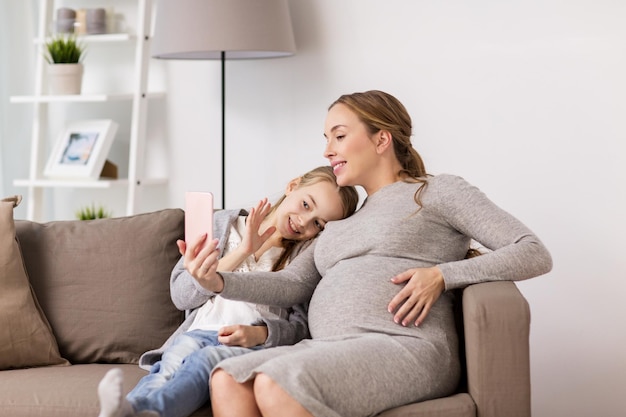 zwangerschap, mensen en familieconcept - gelukkige zwangere vrouw en meisje zittend op de bank en selfie maken met smartphone thuis