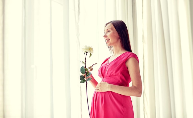 zwangerschap, moederschap, mensen, vakantie en verwachtingsconcept - gelukkige zwangere vrouw met witte roze bloem thuis