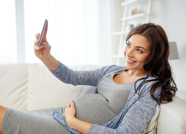 zwangerschap, moederschap, technologie, mensen en verwachtingsconcept - gelukkige zwangere vrouw met smartphone die selfie thuis neemt