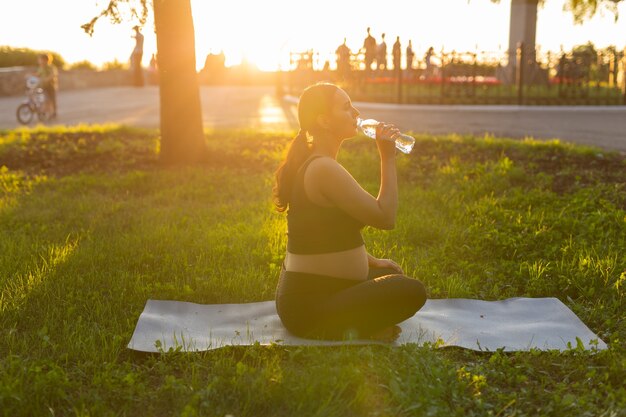 Zwangerschap, sport en gezondheid levensstijl - zwangere vrouw drinkwater uit een fles zittend op het gras in zonnige zomerdag na yoga