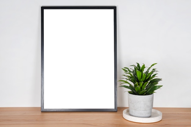 Zwart houten verticaal frame met witte lege kaart en groene plant in betonnen pot op houten tafel op lichtgrijze muur