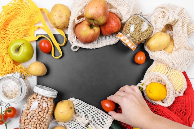 Zwart schoolbord en verschillende groenten in een katoenen ecozak, een netzak en een glazen pot met peulvruchten, gebruik eco-verpakkingen in de keuken, kopieer ruimte