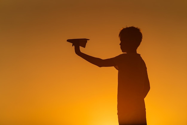 Zwart silhouet van een jongen die tegen een oranje zomerzonsondergang staat met een klein papieren vliegtuigje in handen Horizontale foto