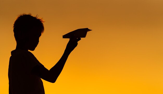 Zwart silhouet van een jongen die tegen een oranje zomerzonsondergang staat met een klein papieren vliegtuigje in handen Horizontale foto