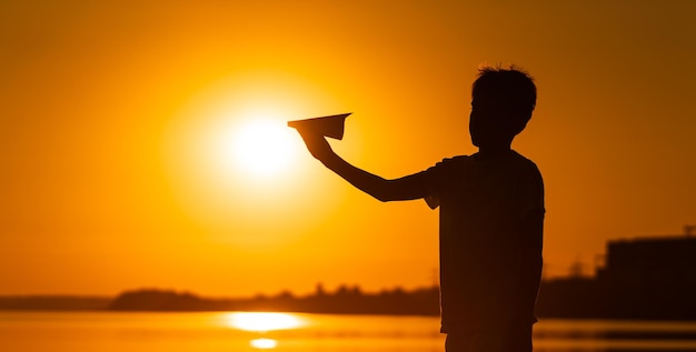 Zwart silhouet van een jongen die tegen een oranje zomerzonsondergang staat met een klein papieren vliegtuigje in handen Horizontale foto