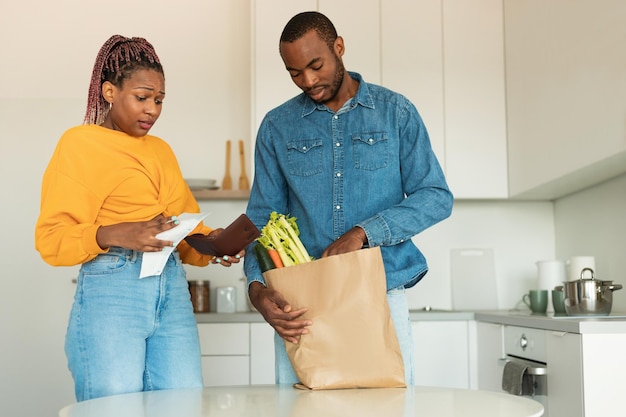 Zwart stel pakt producten uit van de markt in de keuken vrouw kijkt naar rekening en portemonnee met geschokte gezichtsuitdrukking