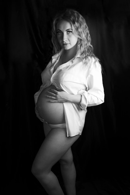 Zwart-wit foto van een zwangere vrouw in de studio op een zwarte achtergrond, staande tot de knie tot de knieën. In een wit overhemd en ondergoed, handen op een blote buik. Een knie gebogen. Kijken naar de camera