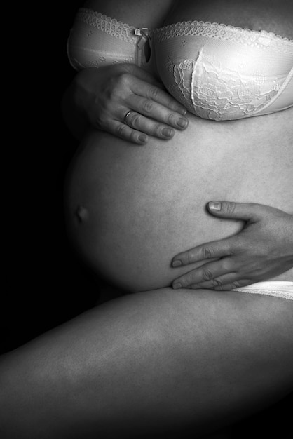 Zwart-wit foto van een zwangere vrouw in wit ondergoed in de studio, op een zwarte achtergrond. op de foto zijn alleen de borsten en een grote ronde buik zichtbaar, met een gebogen been. handen op de buik