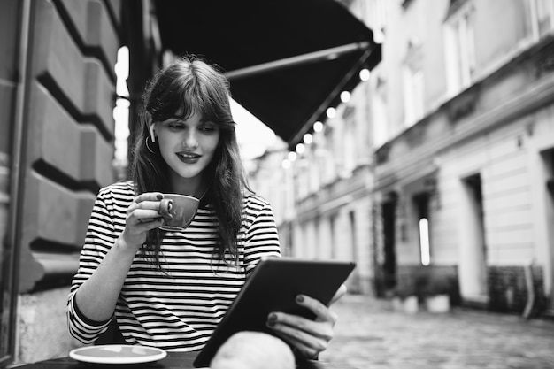 Zwart-wit foto van vrouw die koffie drinkt met digitale tablet en koptelefoon op caféterras