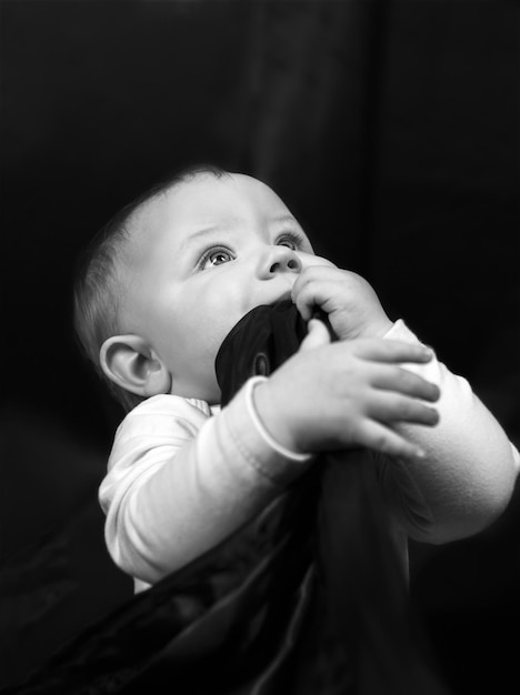 Zwart-wit portret van een eenjarige baby Het kind zit op een zwarte stoffen achtergrond met zijn hoofd omhoog en kijkt omhoog met één hand die de achtergrondstof in zijn mond stopt