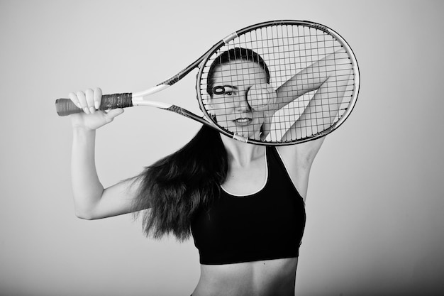 Zwart-wit portret van een mooie jonge vrouw die in sportkleding een tennisracket vasthoudt terwijl ze tegen een witte achtergrond staat