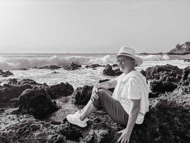 Zwart-wit portret van glimlachende oudere vrouw die op een rotsachtig strand zit en naar de golven van de oceaan kijkt