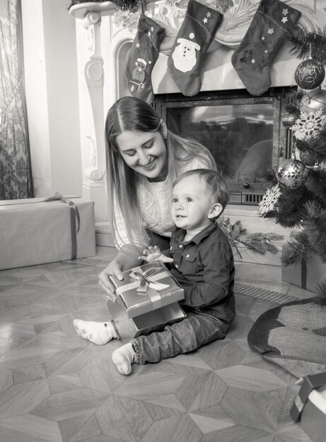 Zwart-witfoto van een gelukkige jonge moeder en een 1-jarige babyjongen op de vloer onder de kerstboom in de woonkamer