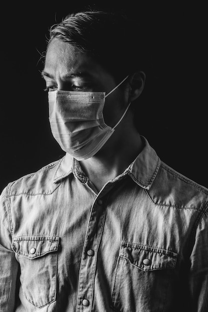 Zwart-witfotografie van een latijnse man die ontmoedigd naar beneden kijkt met een medisch masker en een blauw shirt op een zwarte achtergrond. effecten van het coronavirus