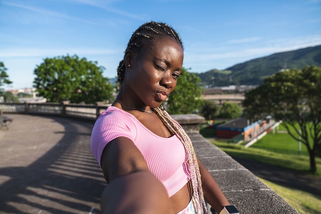 Zwarte Afrikaanse vrouw die een selfie neemt met de telefoon met haar handen erg blij en een kus geeft in een openbaar park op een zeer zonnige dag en grijze lucht. Zwarte vrouw in casual lifestyle kleding