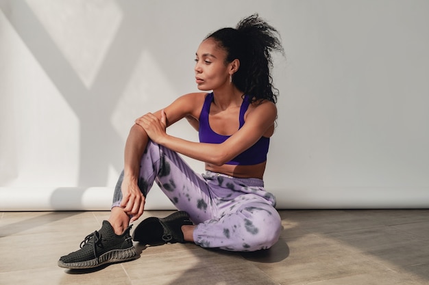 zwarte afro-amerikaanse vrouw zit in een stijlvolle hipster fitness outfit violet top en broek op wit