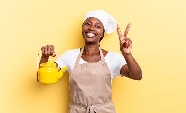 Zwarte afro chef-kok vrouw lacht en ziet er gelukkig, zorgeloos en positief uit, gebarend overwinning of vrede met één hand. theepot concept