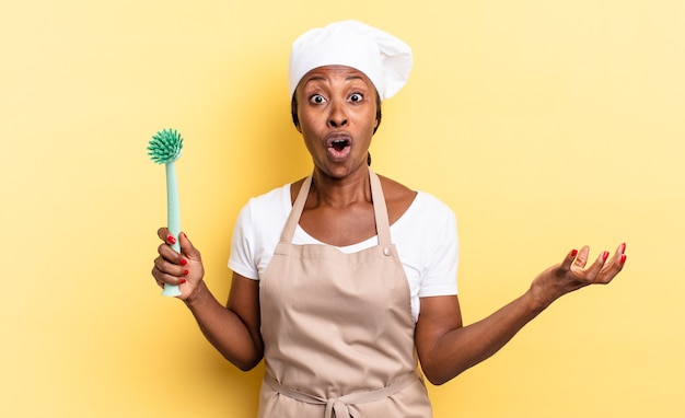 Zwarte afro chef-kok vrouw met open mond en verbaasd, geschokt en verbaasd met een ongelooflijke verrassing. afwas concept