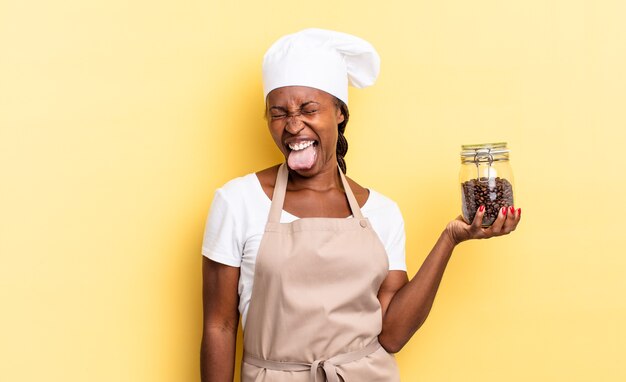 Zwarte afro chef-kok vrouw met vrolijke, zorgeloze, rebelse houding, grappen maken en tong uitsteken, plezier maken. koffiebonen concept