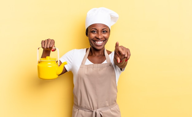 Zwarte afro chef-kok vrouw wijzend op camera met een tevreden, zelfverzekerde, vriendelijke glimlach, jou kiezen. theepot concept