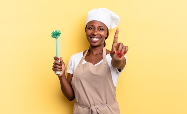 Zwarte afro-chef-vrouw die trots en zelfverzekerd glimlacht en nummer één poseert, triomfantelijk en zich een leider voelt als een concept voor het schoonmaken van gerechten
