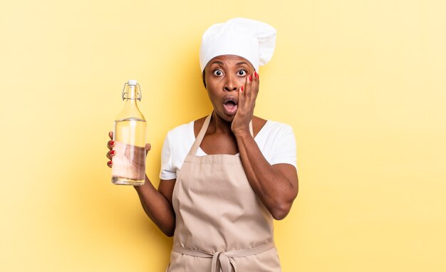 zwarte afro-chef-vrouw die zich geschokt en bang voelt, er doodsbang uitziet met open mond en handen op de wangen met een waterfles