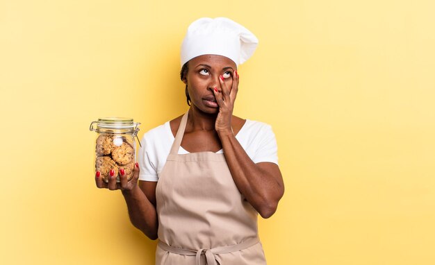 Zwarte afro-chef-vrouw die zich verveeld, gefrustreerd en slaperig voelt na een vermoeiende, saaie en vervelende taak, gezicht met de hand vasthoudend. koekjes concept