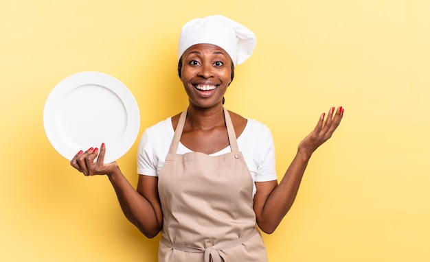 Zwarte afro-chef-vrouw voelt zich gelukkig, verrast en opgewekt, glimlacht met een positieve houding en realiseert een oplossing of idee. leeg bord concept