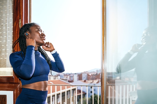 Zwarte afrovrouw gekleed in sportkleding die naar muziek luistert met een koptelefoon in een raam van haar huis omdat ze thuis gaat sporten vanwege de covid19 coronavirus pandemie