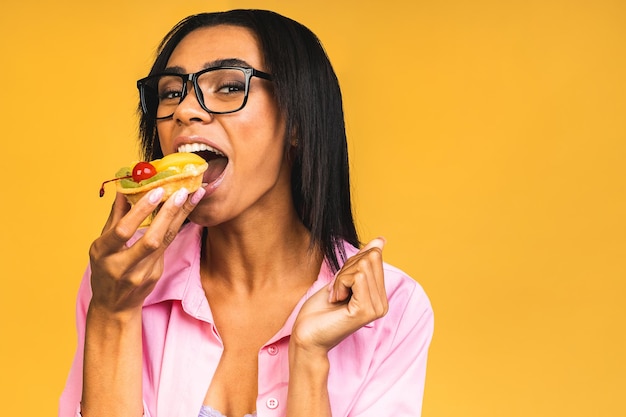 Zwarte Amerikaanse Afrikaanse gelukkige vrouw die cakedessert eet die over gele achtergrond wordt geïsoleerd Het eten van cupcake Dieet ongezond voedselconcept