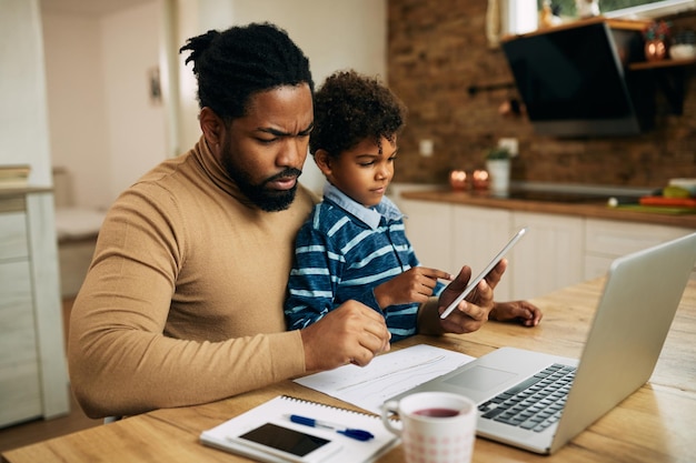 Zwarte blijf thuis vader werkt op laptop terwijl zijn kleine zoon thuis touchpad gebruikt