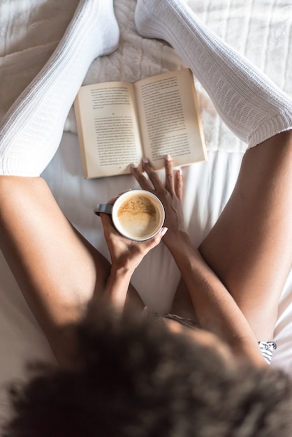 Foto zwarte die een boek leest en koffie op bed met sokken drinkt