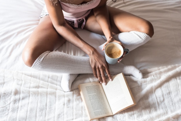 Foto zwarte die een boek leest en koffie op bed met sokken drinkt