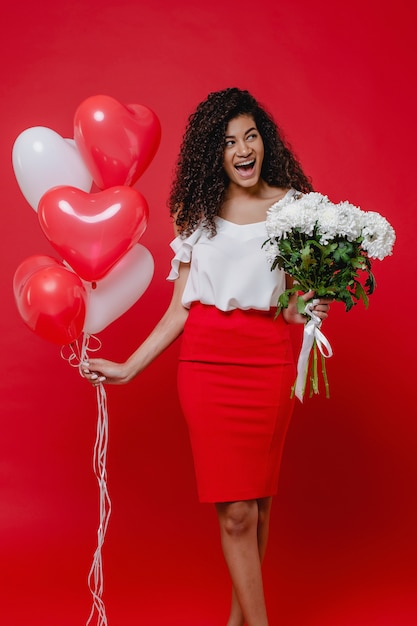 Zwarte die met hart gevormde ballons en boeket van witte bloemen op rode muur glimlacht